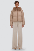 A model stands against a light gray studio background wearing a cropped beige puffer jacket with a large brown faux-fur collar and wide-leg beige trousers.