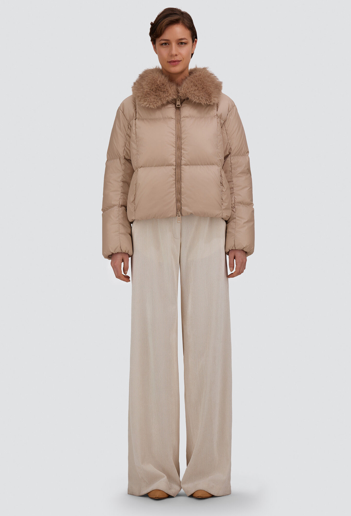 A model stands against a light gray studio background wearing a cropped beige puffer jacket with a large brown faux-fur collar and wide-leg beige trousers.