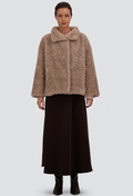 A model stands against a light gray studio background wearing a cropped, shaggy faux-fur jacket in a warm taupe-brown color, paired with a long dark brown skirt and black ankle boots.