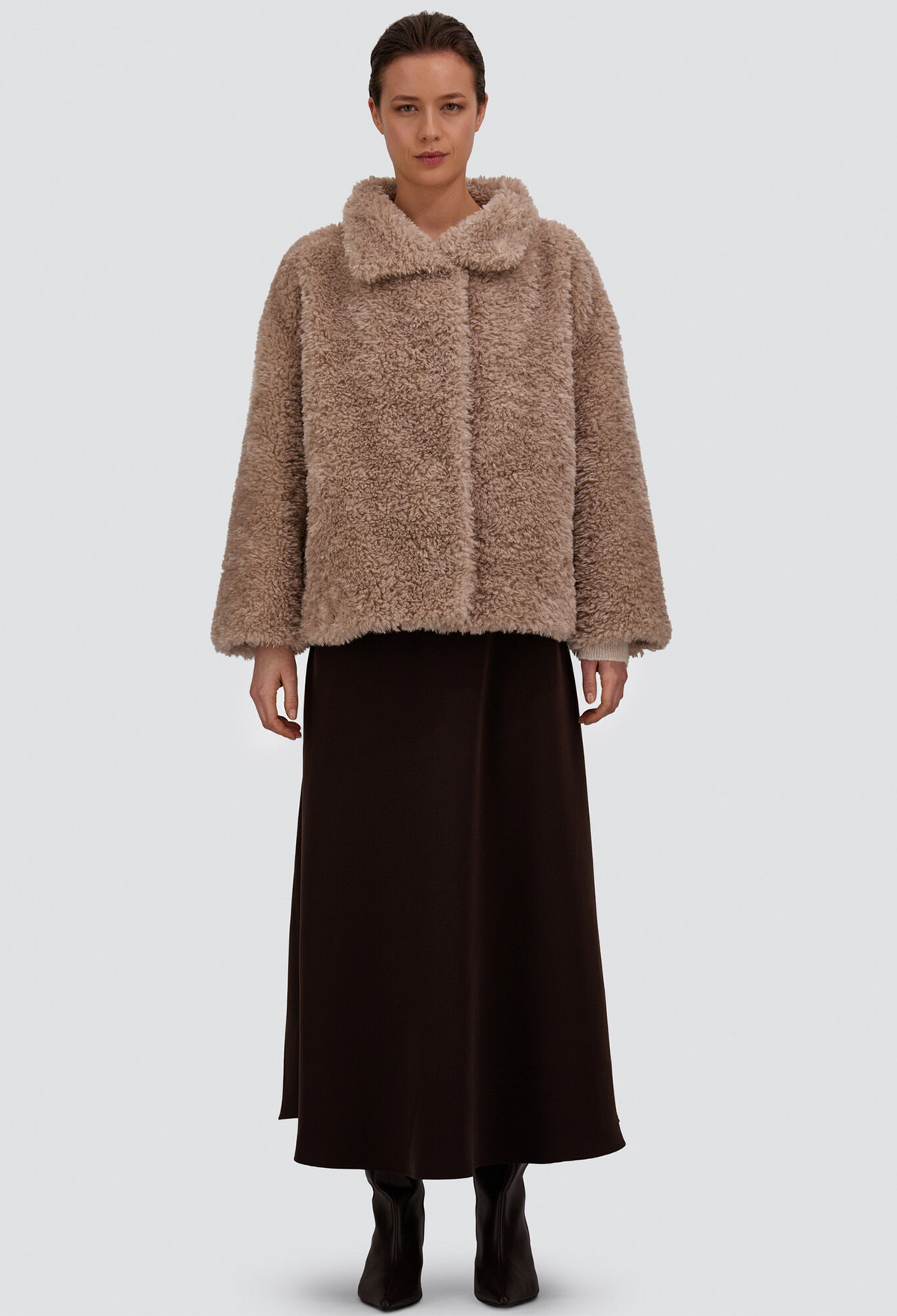 A model stands against a light gray studio background wearing a cropped, shaggy faux-fur jacket in a warm taupe-brown color, paired with a long dark brown skirt and black ankle boots.