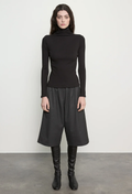 A model stands against a light gray wall wearing a black long-sleeve mock-neck top, black draped wide-leg skirt, and black knee-high boots.