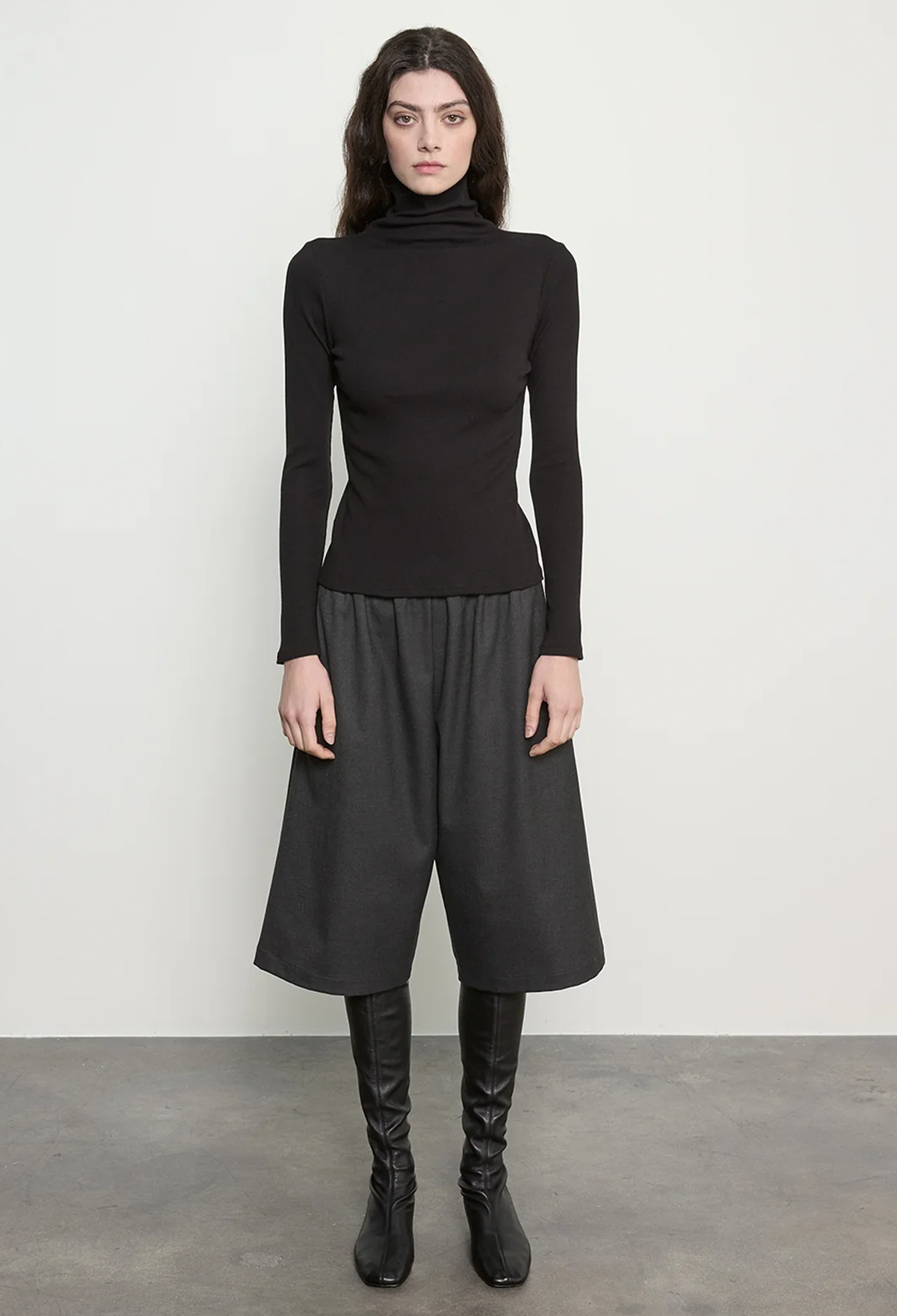 A model stands against a light gray wall wearing a black long-sleeve mock-neck top, black draped wide-leg skirt, and black knee-high boots.
