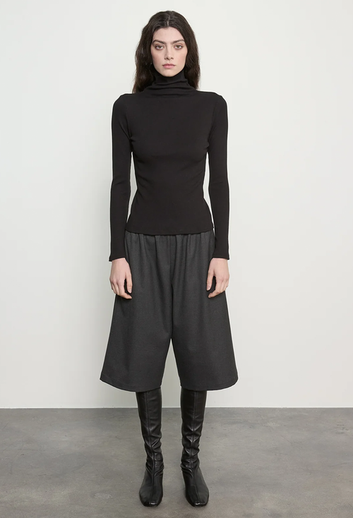 A model stands against a light gray wall wearing a black long-sleeve mock-neck top, black draped wide-leg skirt, and black knee-high boots.