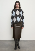 A model stands against a plain light gray wall wearing a black, light blue, and white argyle sweater, a dark brown leather skirt, and black knee-high boots.