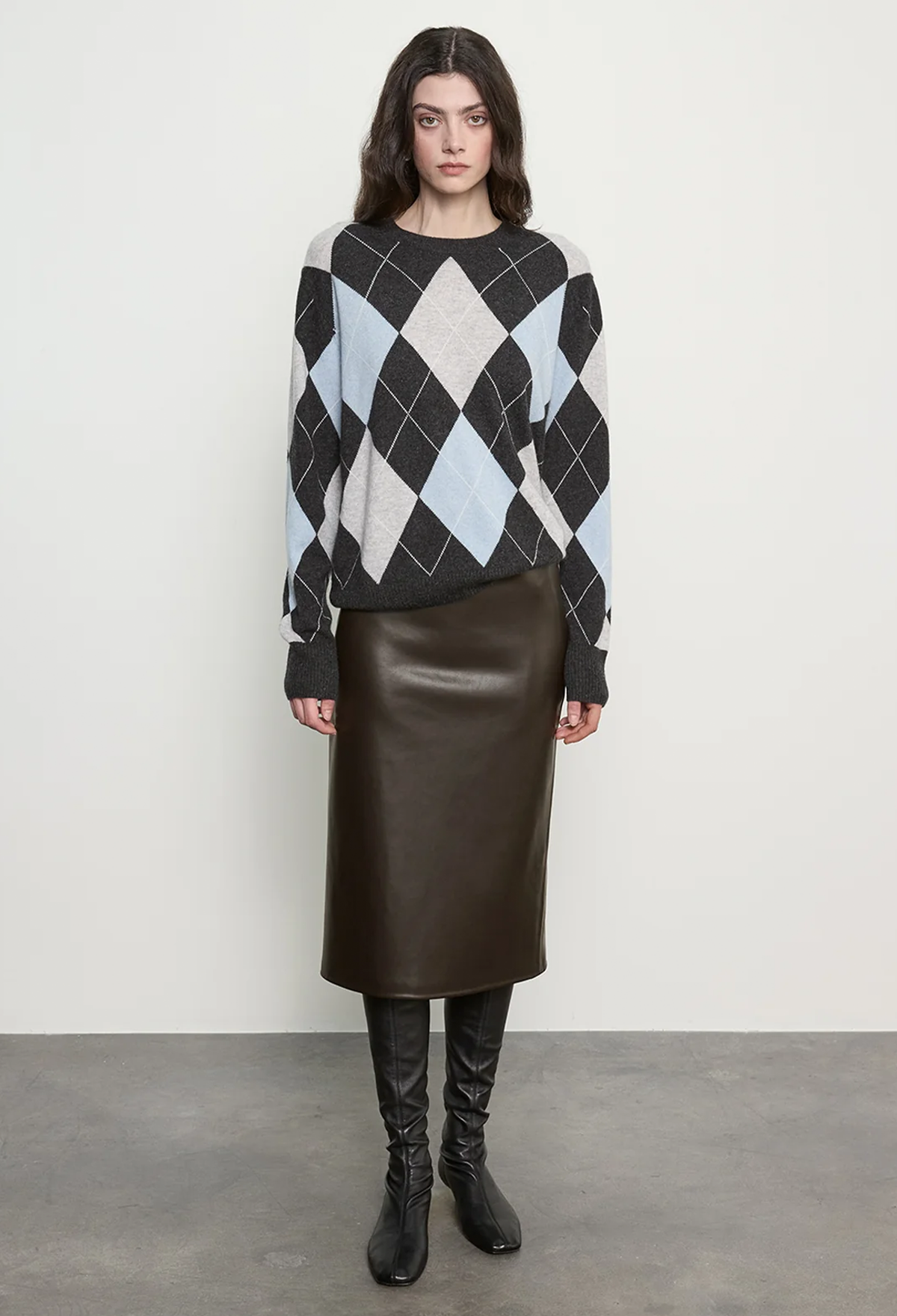 A model stands against a plain light gray wall wearing a black, light blue, and white argyle sweater, a dark brown leather skirt, and black knee-high boots.