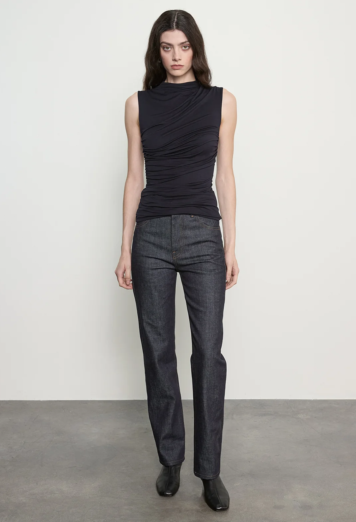 A model stands against a plain light gray studio wall wearing a sleeveless black ruched top, dark charcoal gray jeans, and black boots.