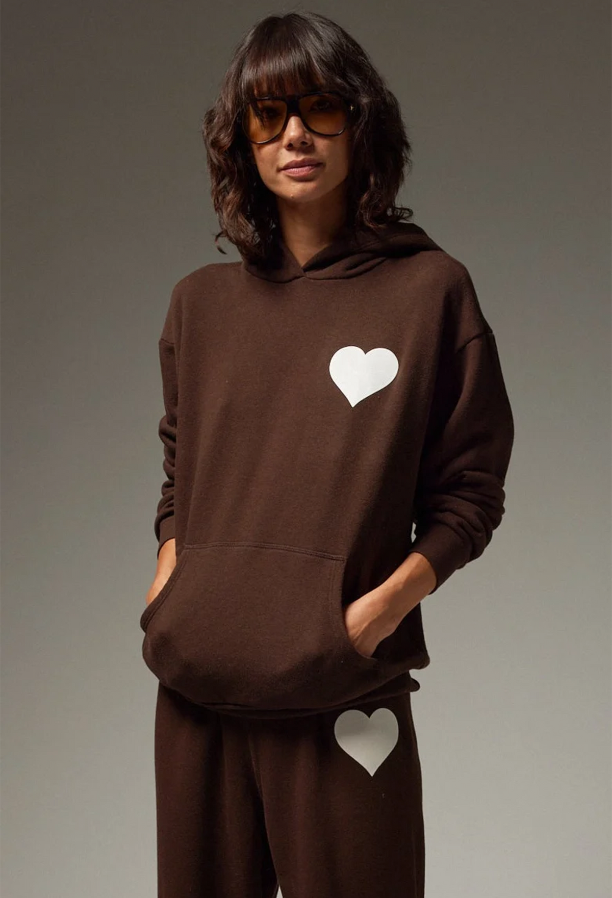 A woman modeling a dark brown matching loungewear set consisting of a pullover hoodie and sweatpants, both featuring a white heart graphic on the chest and thigh, set against a neutral gray studio background.