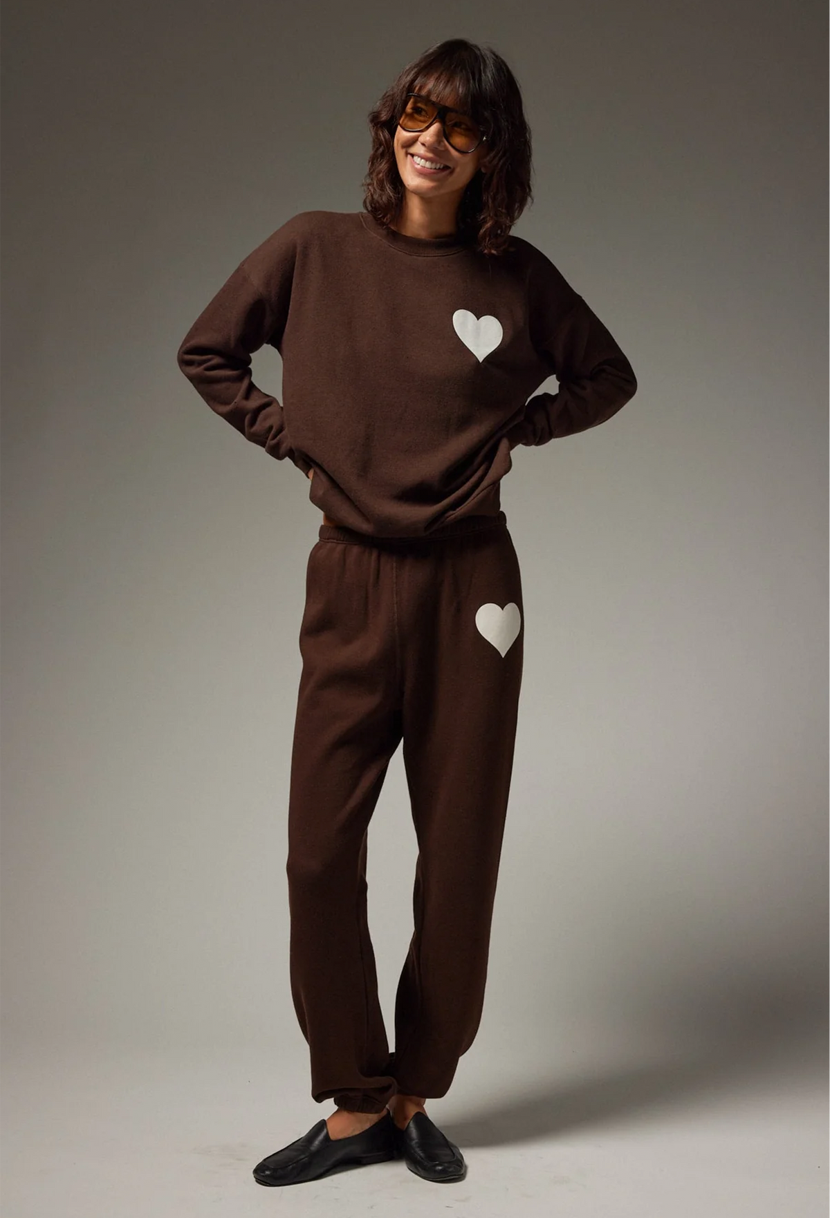A woman modeling a matching brown sweatshirt and jogger pants set with white heart graphics, styled with black slip-on shoes against a neutral gray studio background.