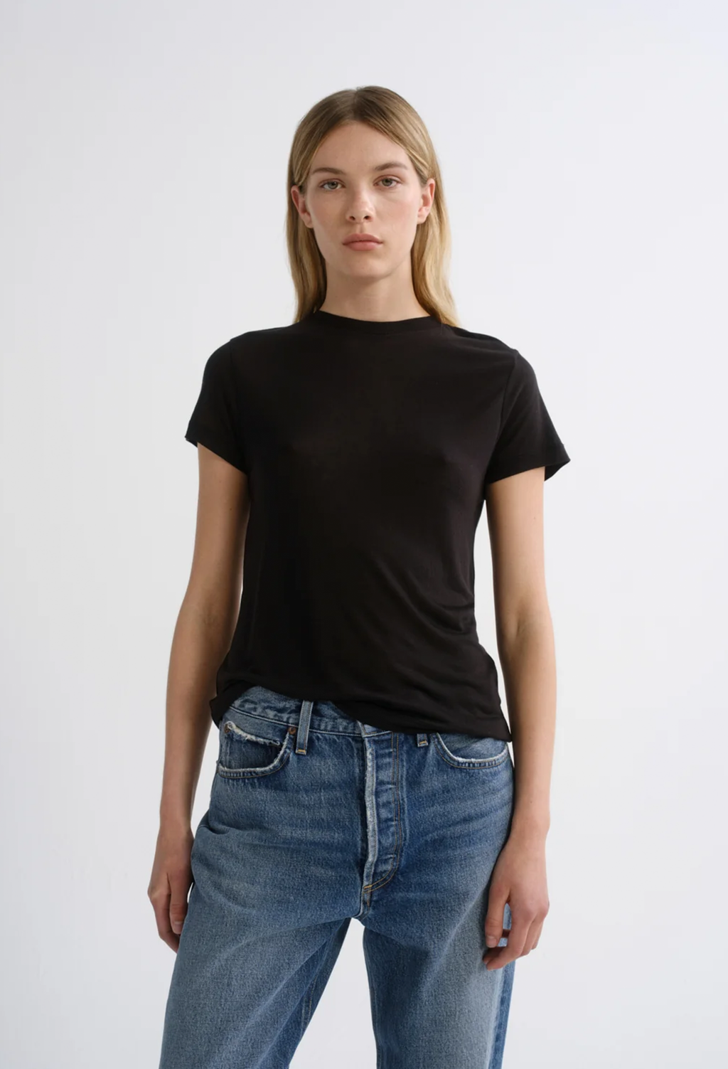 A woman modeling a black short-sleeve crewneck t-shirt paired with blue jeans, standing against a plain light gray studio background.