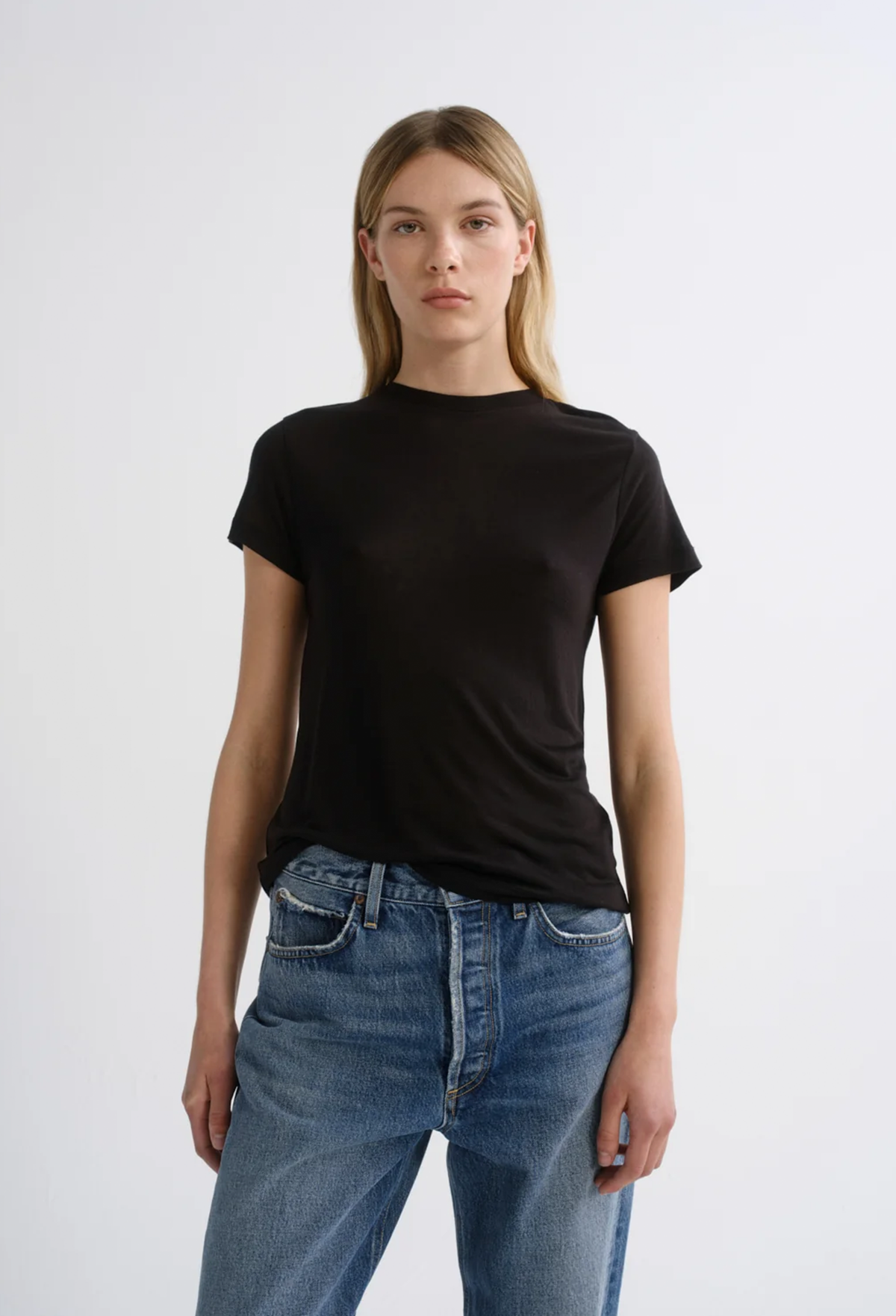 A woman modeling a black short-sleeve crewneck t-shirt paired with blue jeans, standing against a plain light gray studio background.