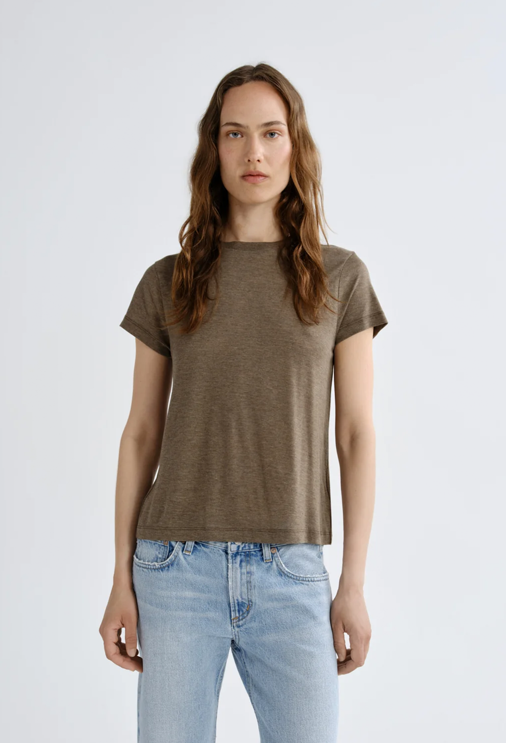 A woman modeling a short-sleeve crewneck t-shirt in a muted brownish-gray color, paired with light-wash blue jeans, against a plain light gray studio background.