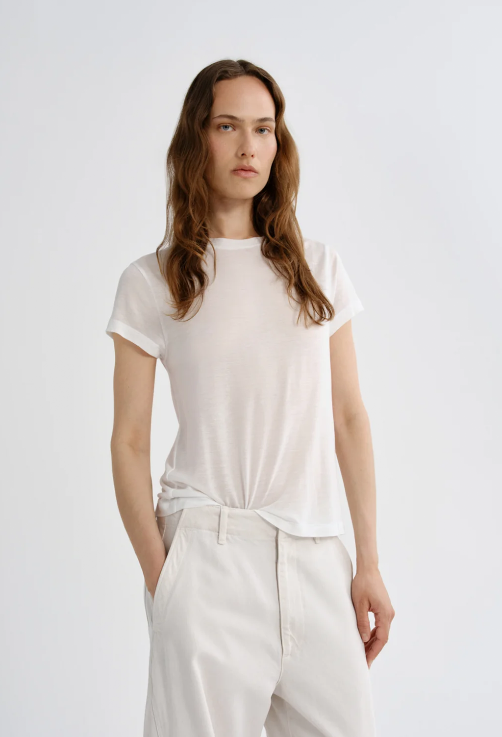 A woman modeling a white short-sleeve crewneck t-shirt tucked into white trousers, photographed against a seamless light gray studio background.