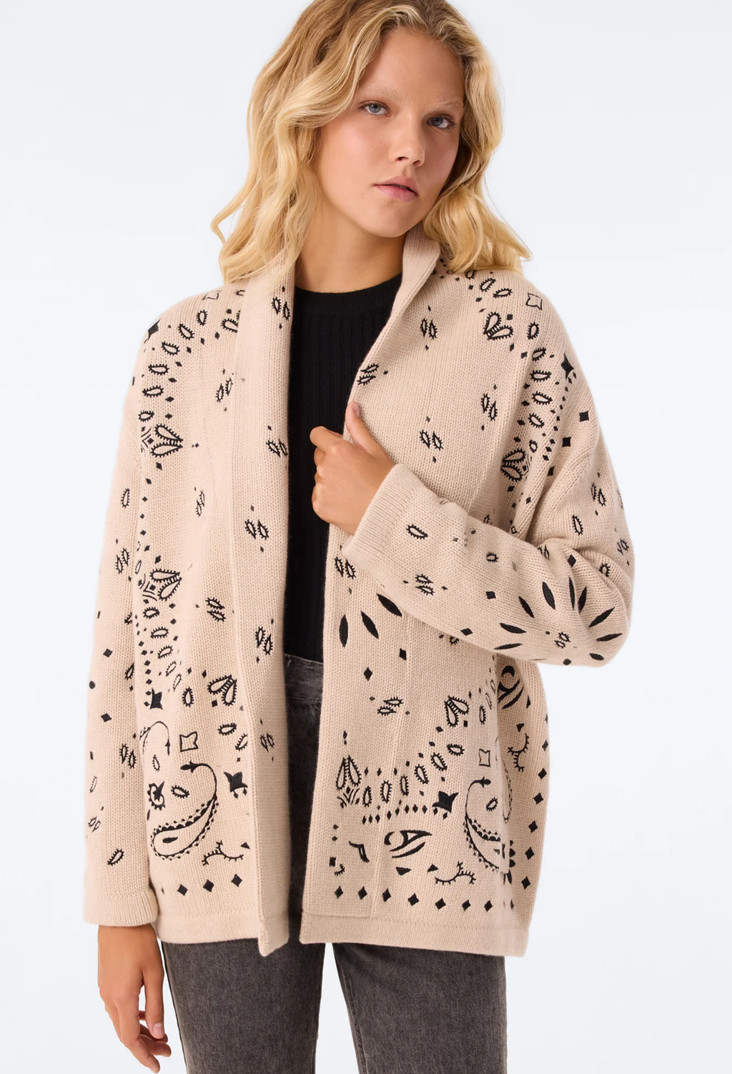 A woman wearing a beige open-front cardigan with black hand-drawn doodle patterns, styled over a black top and gray pants, against a plain white background.