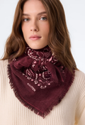 A woman wearing a deep burgundy bandana scarf with a paisley pattern and fringe. She is wearing a cream-colored ribbed sweater against a plain white background.