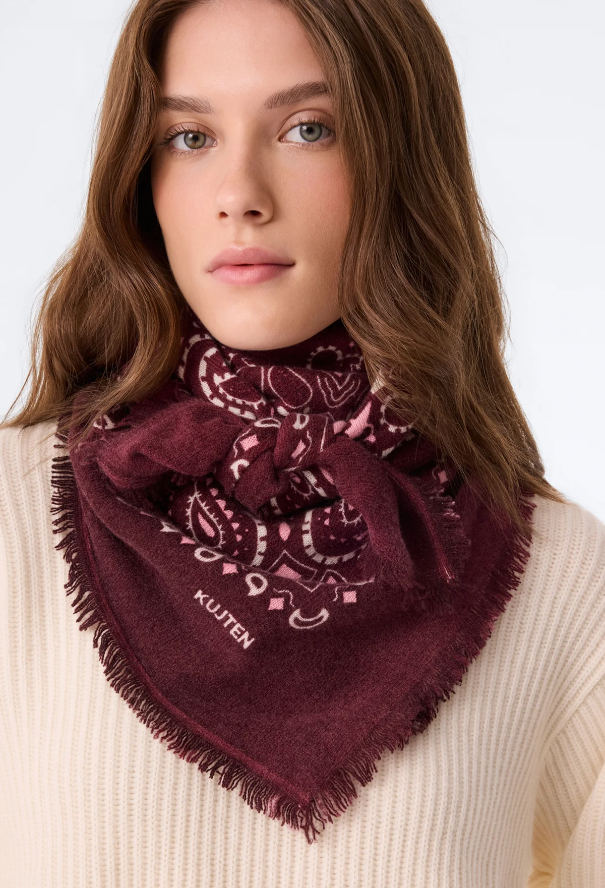 A woman wearing a deep burgundy bandana scarf with a paisley pattern and fringe. She is wearing a cream-colored ribbed sweater against a plain white background.
