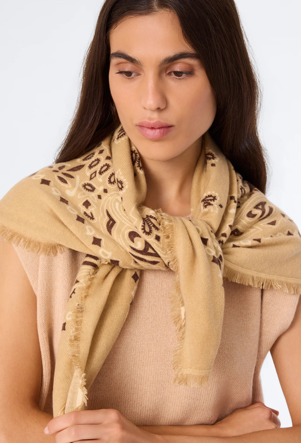 A woman wearing a beige scarf with a brown abstract pattern and fringed edges, styled over a beige top against a light gray background.