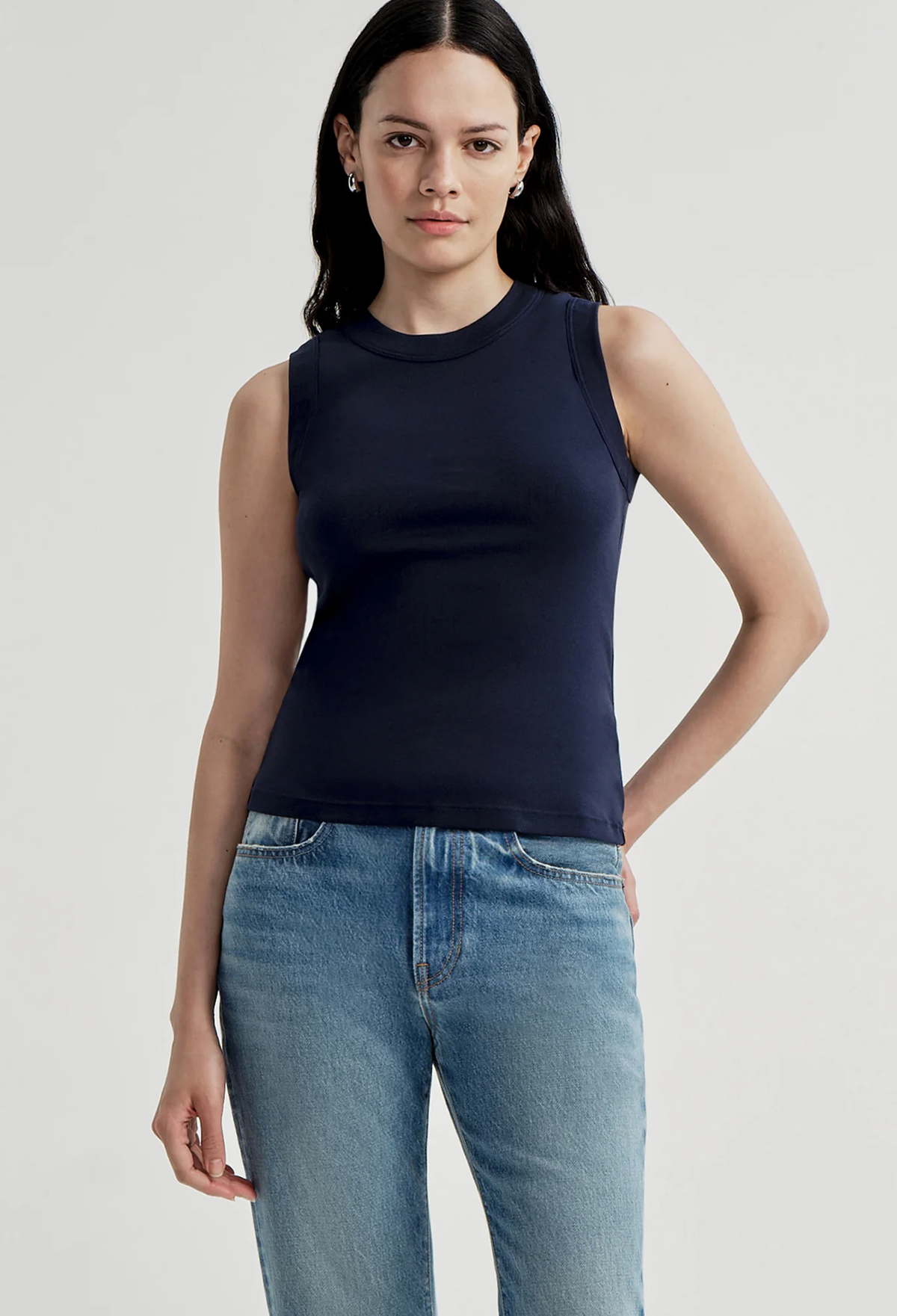 A woman modeling a sleeveless navy blue mock neck top paired with light wash blue jeans, photographed against a neutral light gray studio background.