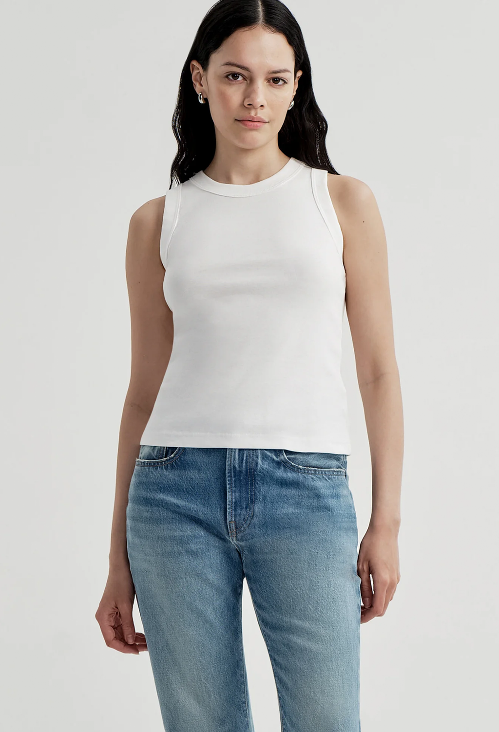 A woman modeling a white ribbed sleeveless crop top paired with blue jeans, photographed against a clean light gray studio background.
