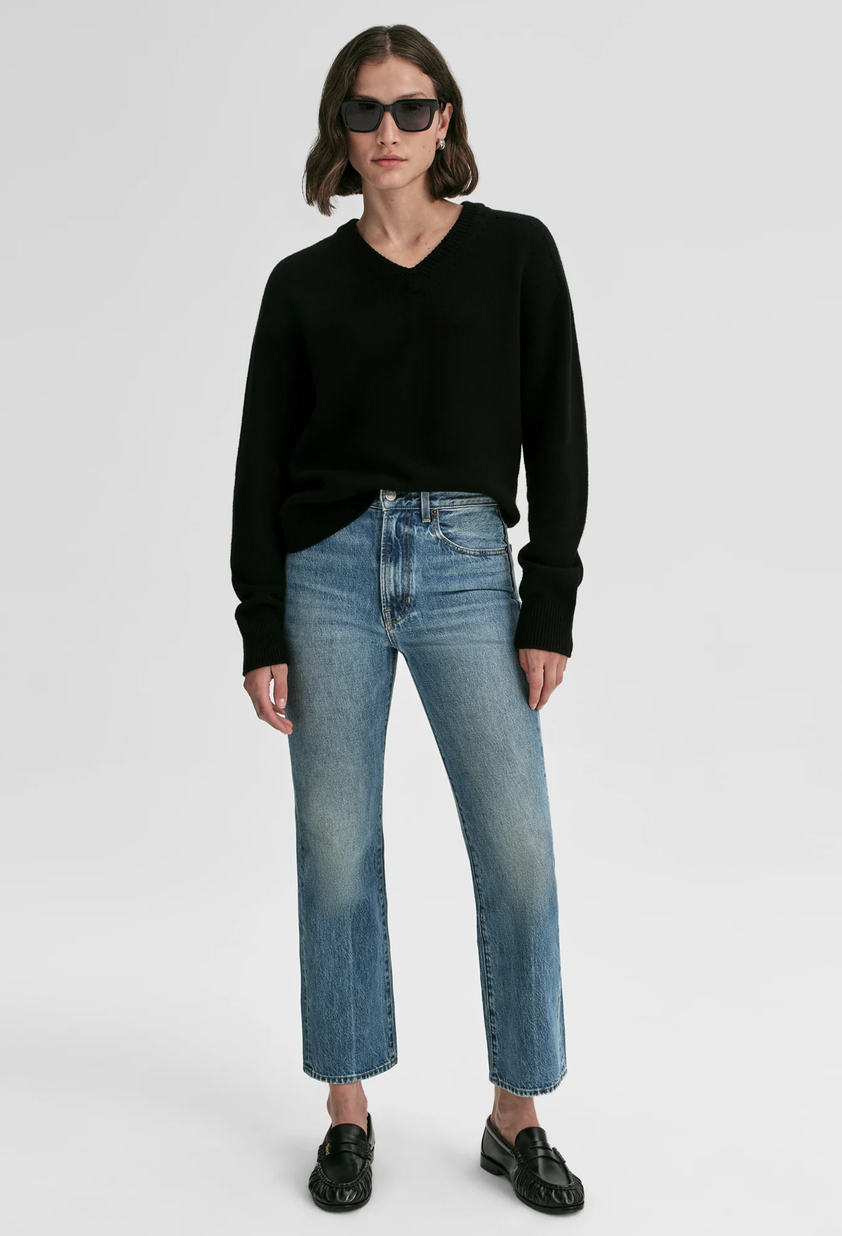 A model wearing a black long-sleeve V-neck sweater, blue cropped jeans with a subtle gradient fade, black loafers, and sunglasses, standing against a light gray studio background.
