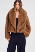 A woman modeling a cropped, long-shawl collar faux fur jacket in a warm camel brown color, paired with dark trousers, against a light gray studio background.