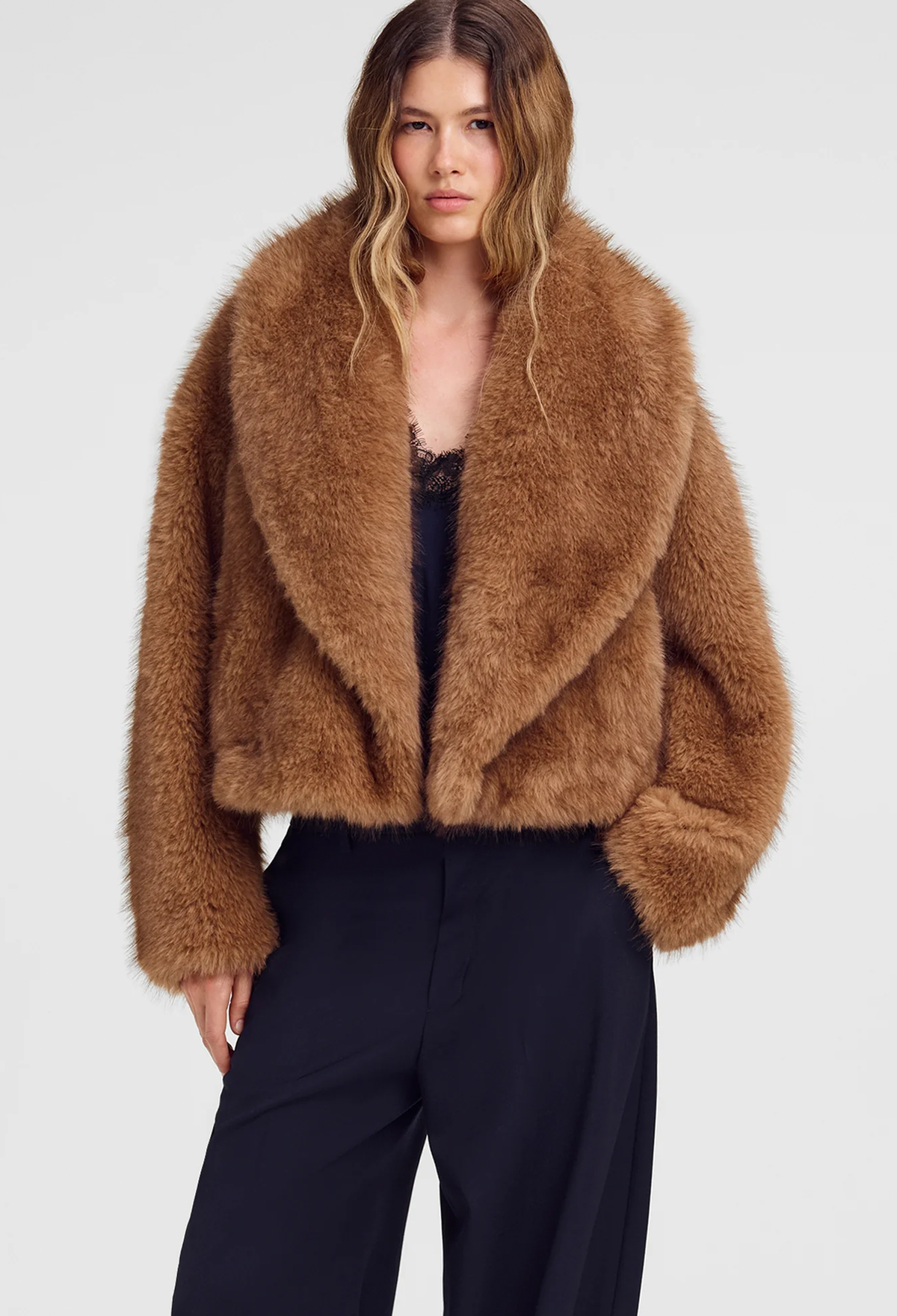 A woman modeling a cropped, long-shawl collar faux fur jacket in a warm camel brown color, paired with dark trousers, against a light gray studio background.