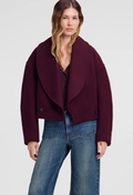 A woman modeling a cropped, oversized deep burgundy wool-blend jacket with a wide shawl collar, paired with blue jeans, against a light gray studio background.