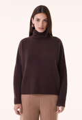 A woman modeling a dark brown ribbed knit turtleneck sweater with matching brown trousers, photographed against a clean off-white studio background.