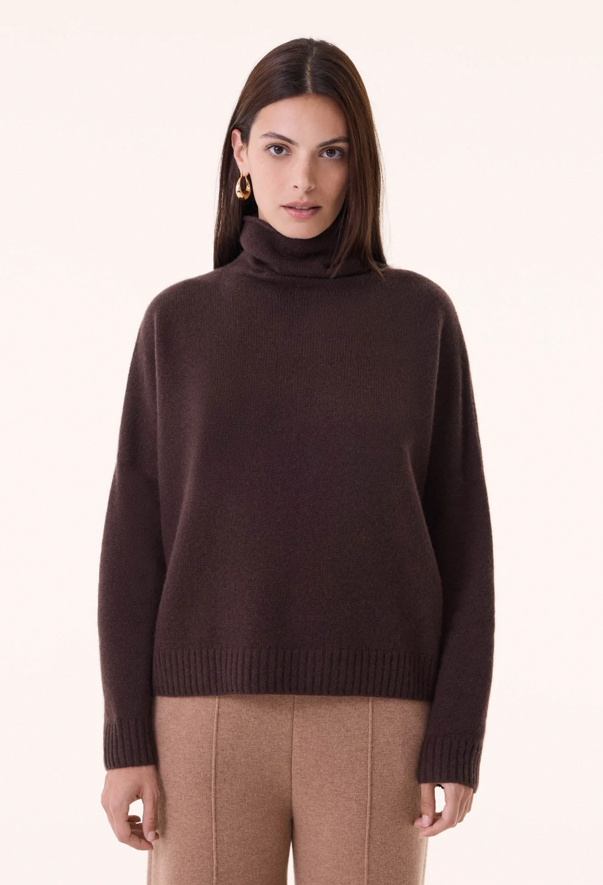 A woman modeling a dark brown ribbed knit turtleneck sweater with matching brown trousers, photographed against a clean off-white studio background.
