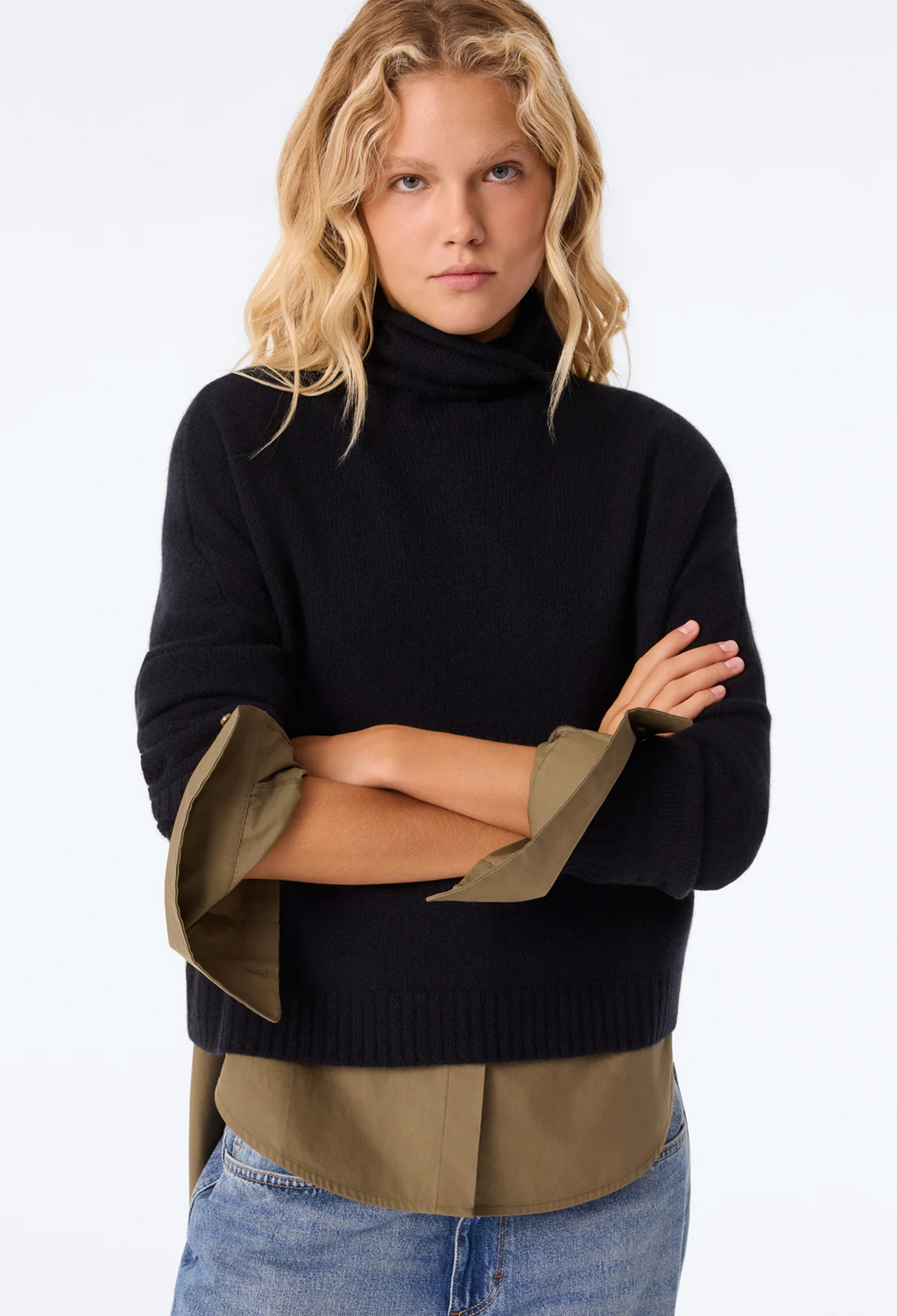 A blonde model wearing a black ribbed turtleneck sweater with wide, turned-up olive green shirt sleeves, layered over a matching olive green shirt and blue jeans, against a plain white studio background.