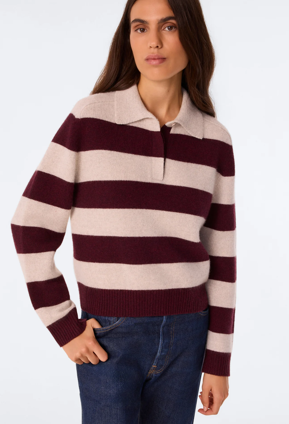 A woman wearing a long-sleeve knit polo-style sweater with horizontal burgundy and cream stripes, paired with blue jeans, against a plain light gray studio background.
