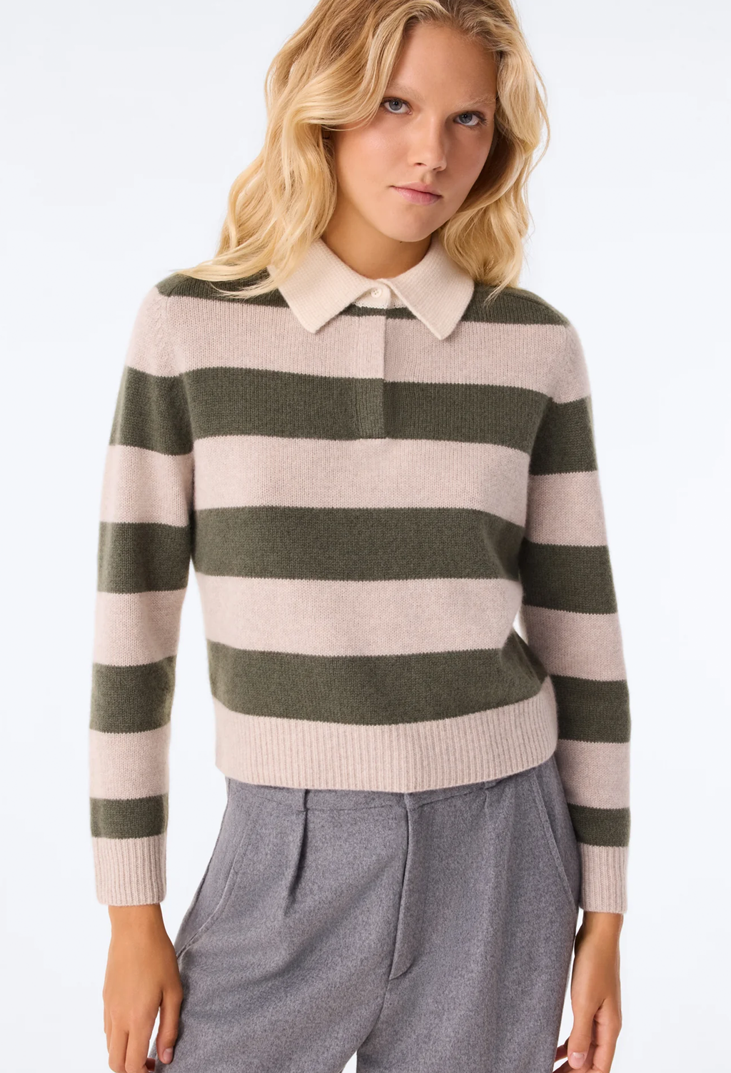 A woman wearing a knit polo-style sweater with wide horizontal stripes in beige and olive green, paired with gray trousers, against a plain white studio background.