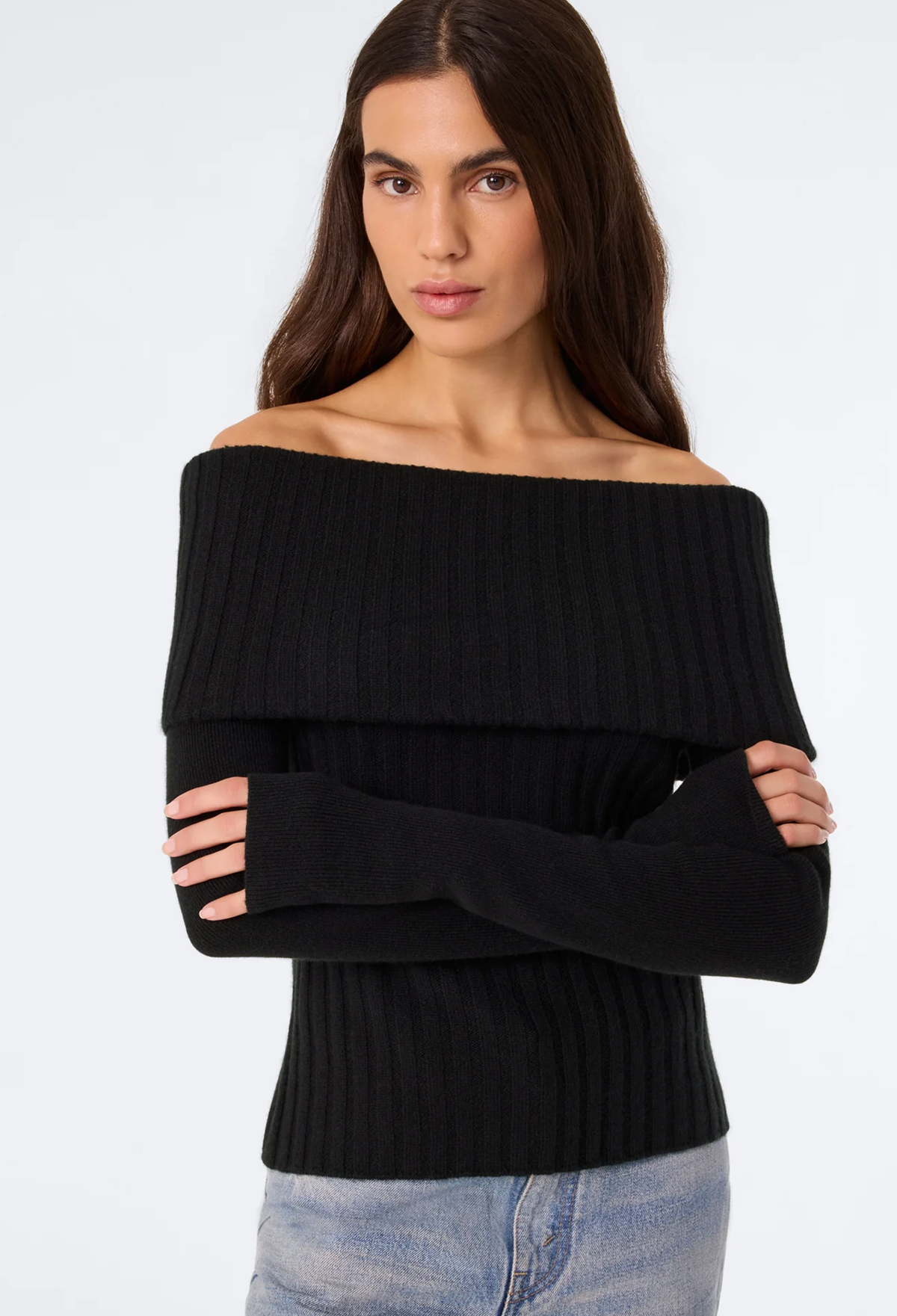 A woman wearing a black off-the-shoulder ribbed knit sweater with long sleeves, paired with light blue jeans, against a plain white studio background.