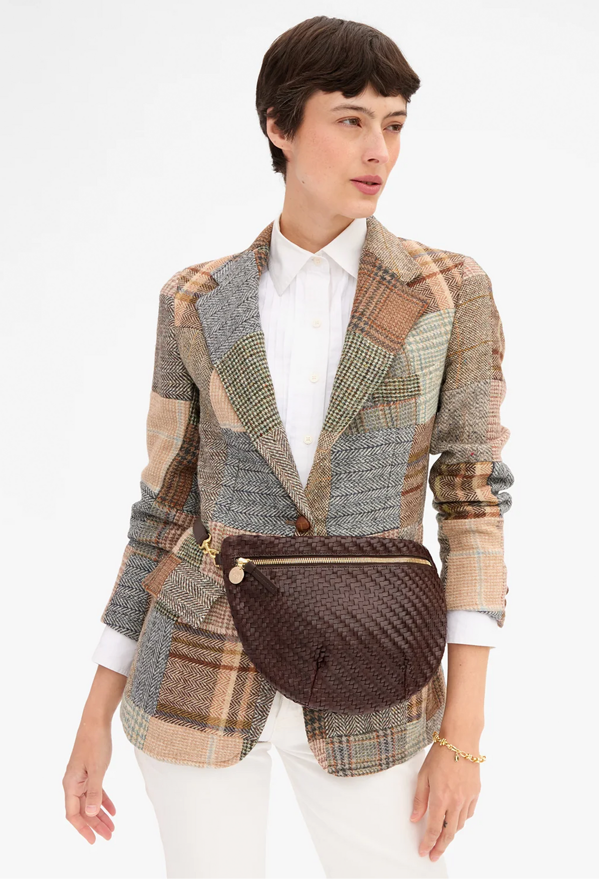 A model wearing a brown woven crossbody belt bag with gold hardware, styled over a multicolor patchwork plaid blazer and white shirt.