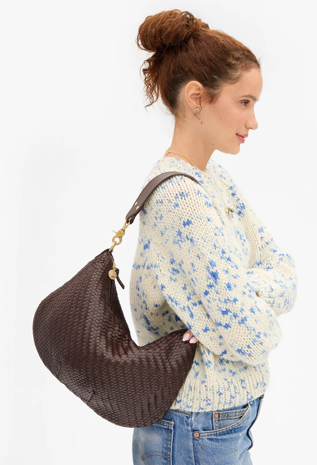 A woman in a cream knit sweater with blue floral patterns and blue jeans carries a large, dark brown woven crescent shoulder bag with gold-tone hardware against a plain white background.