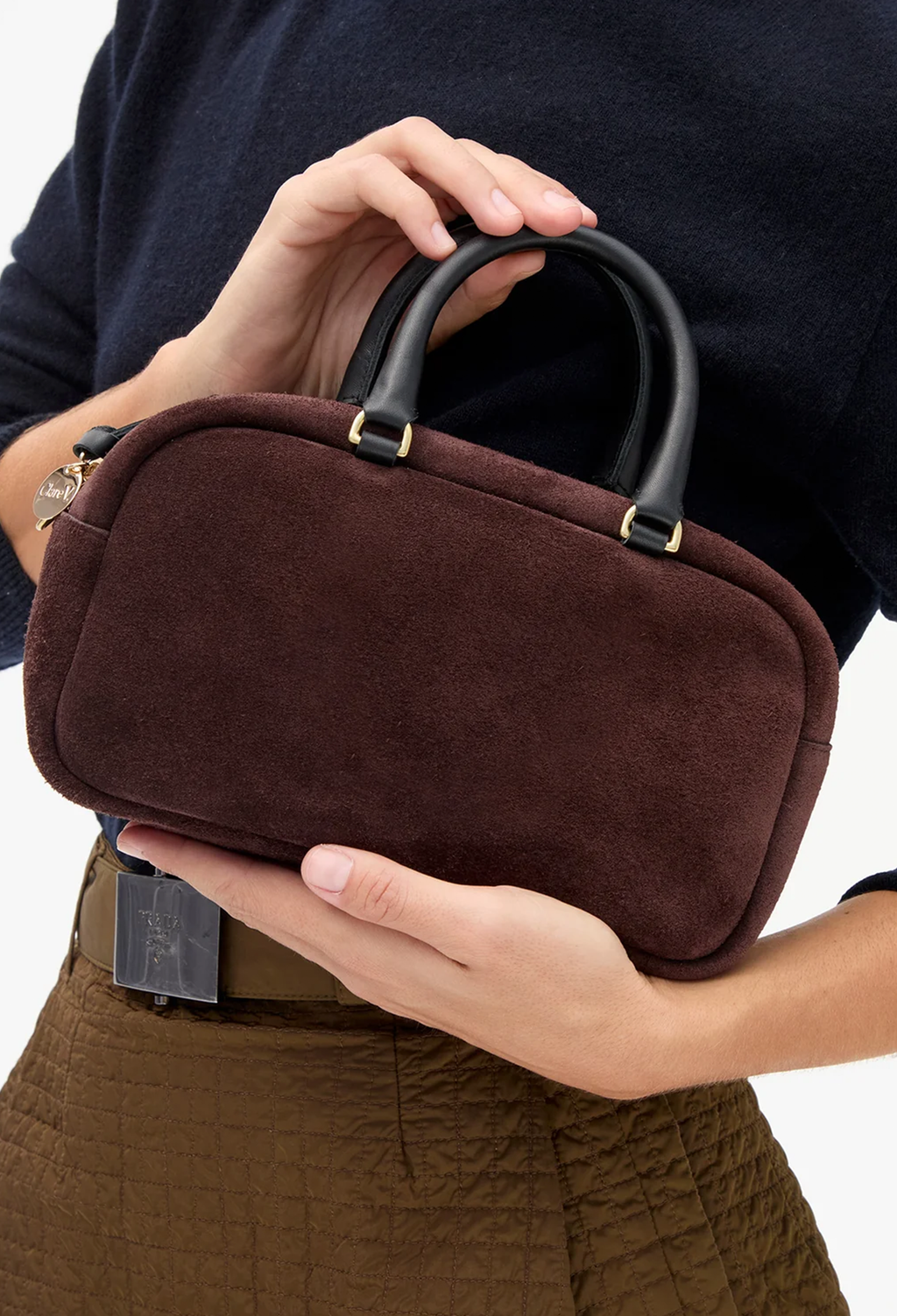 A person holding a small structured handbag in dark brown suede with black leather top handles and gold-tone hardware, shown against a plain light background.