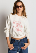 A woman wearing a cream-colored cropped sweatshirt with a pink retro graphic that reads 'Things Go Better With Coke' and blue jeans, posing against a light gray background.