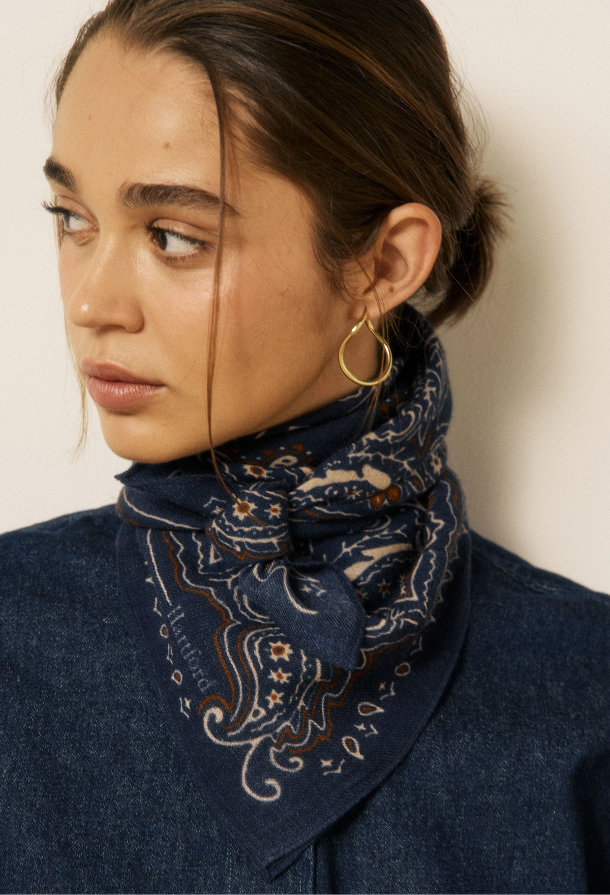 A close-up of a woman wearing a navy blue paisley bandana scarf tied around her neck, paired with a dark denim shirt and gold hoop earrings against a plain light background.
