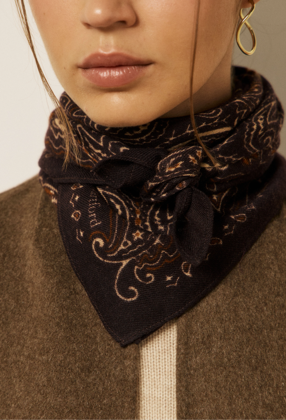 A close-up of a model wearing a dark brown paisley bandana scarf tied around their neck, paired with a brown knit sweater.