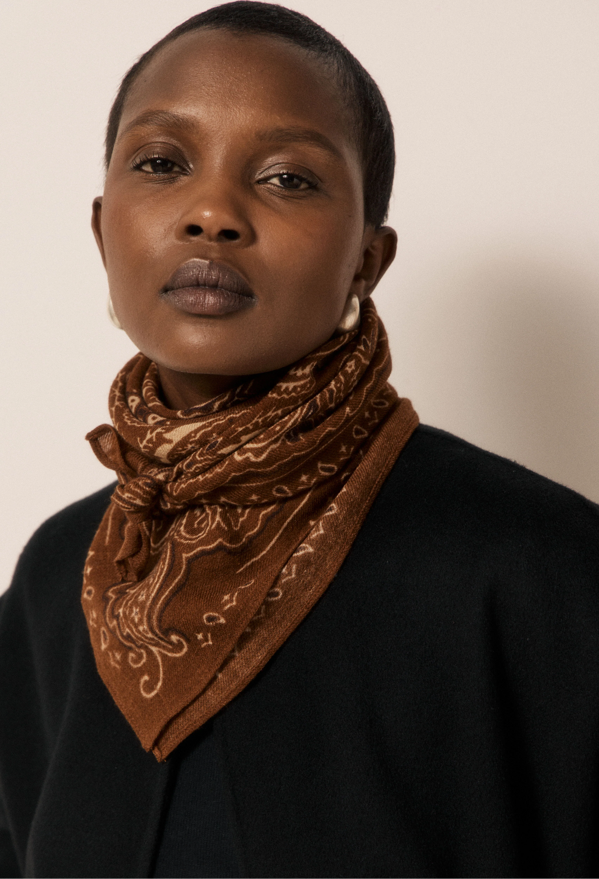 A close-up portrait of a model wearing a brown paisley bandana scarf tied around the neck, paired with a black top against a plain light beige background.