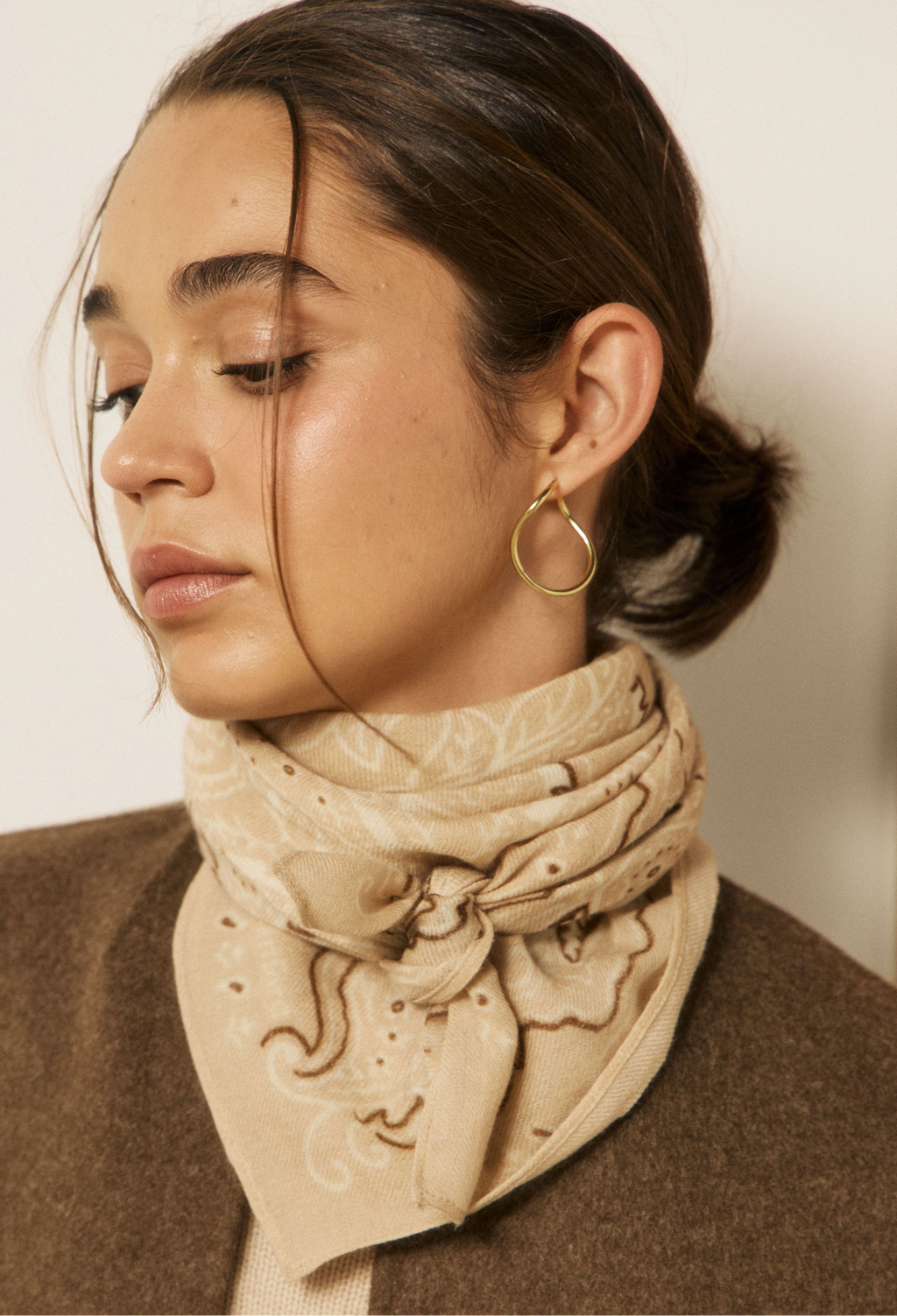 A close-up of a woman wearing a beige patterned bandana scarf and a brown wool sweater, with a gold hoop earring visible.