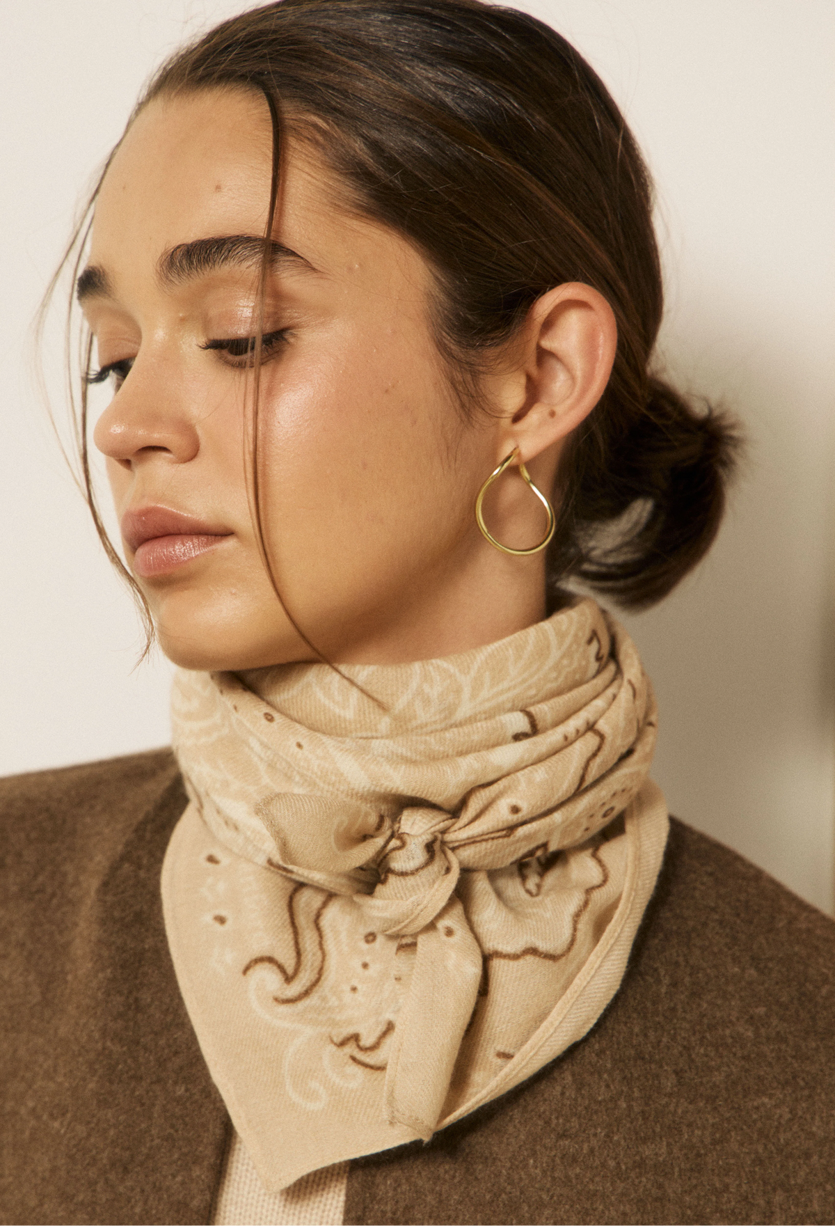 A close-up of a woman wearing a beige patterned bandana scarf and a brown wool sweater, with a gold hoop earring visible.