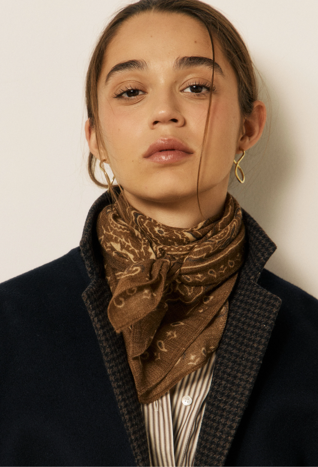A close-up portrait of a woman wearing a brown patterned bandana scarf tied around her neck, a dark navy coat, and a striped shirt. She is wearing gold hoop earrings against a plain off-white background.