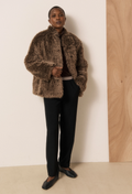 A model stands against a minimalist beige and wood-paneled wall wearing a brown shaggy faux-fur cropped jacket over a dark top and black trousers.