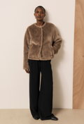 A model wearing a brown fuzzy short coat paired with black wide-leg trousers and black shoes, standing against a minimalist beige and wood-paneled wall.