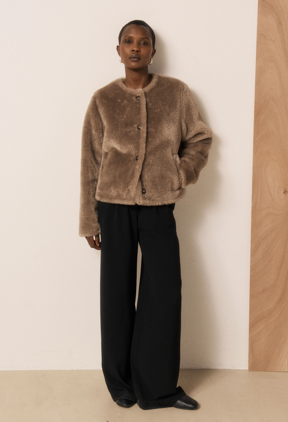 A model wearing a brown fuzzy short coat paired with black wide-leg trousers and black shoes, standing against a minimalist beige and wood-paneled wall.