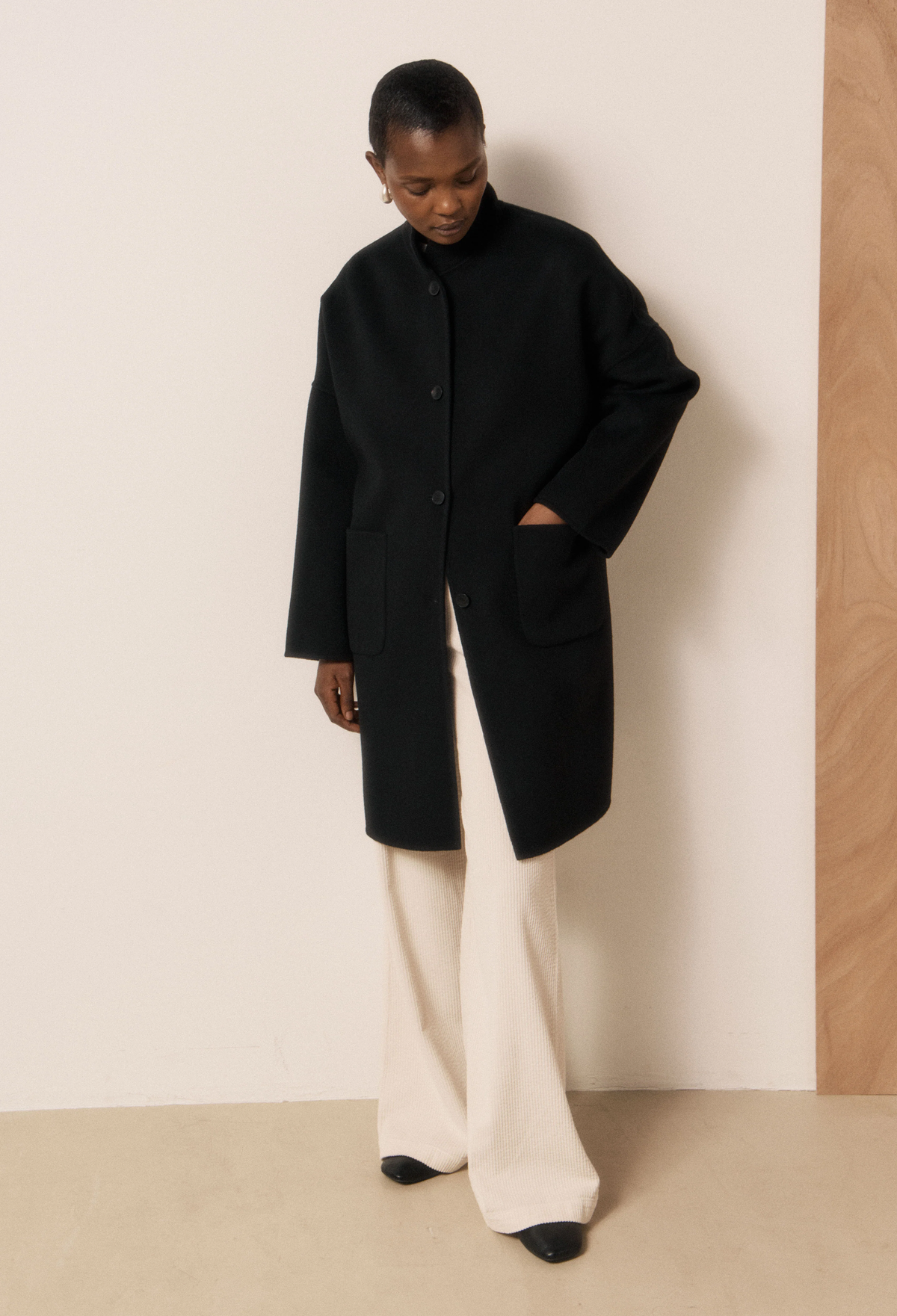 A model wearing a black long-line coat with a high collar, paired with cream wide-leg trousers and black shoes, standing against a minimalist beige and wood-paneled wall.