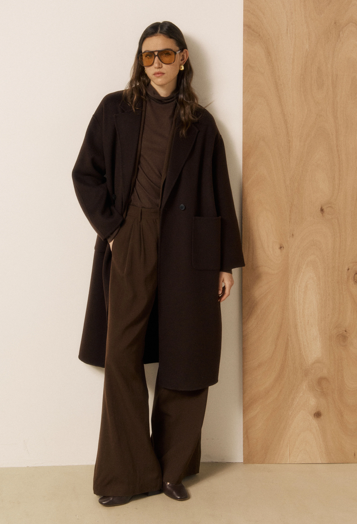 A model wearing a dark brown double-breasted coat, matching brown turtleneck, wide-leg trousers, and sunglasses, standing against a split white and wood-paneled wall.