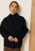 A woman wearing a dark navy oversized blazer over a black turtleneck sweater, standing against a minimalist beige and wood-paneled wall.