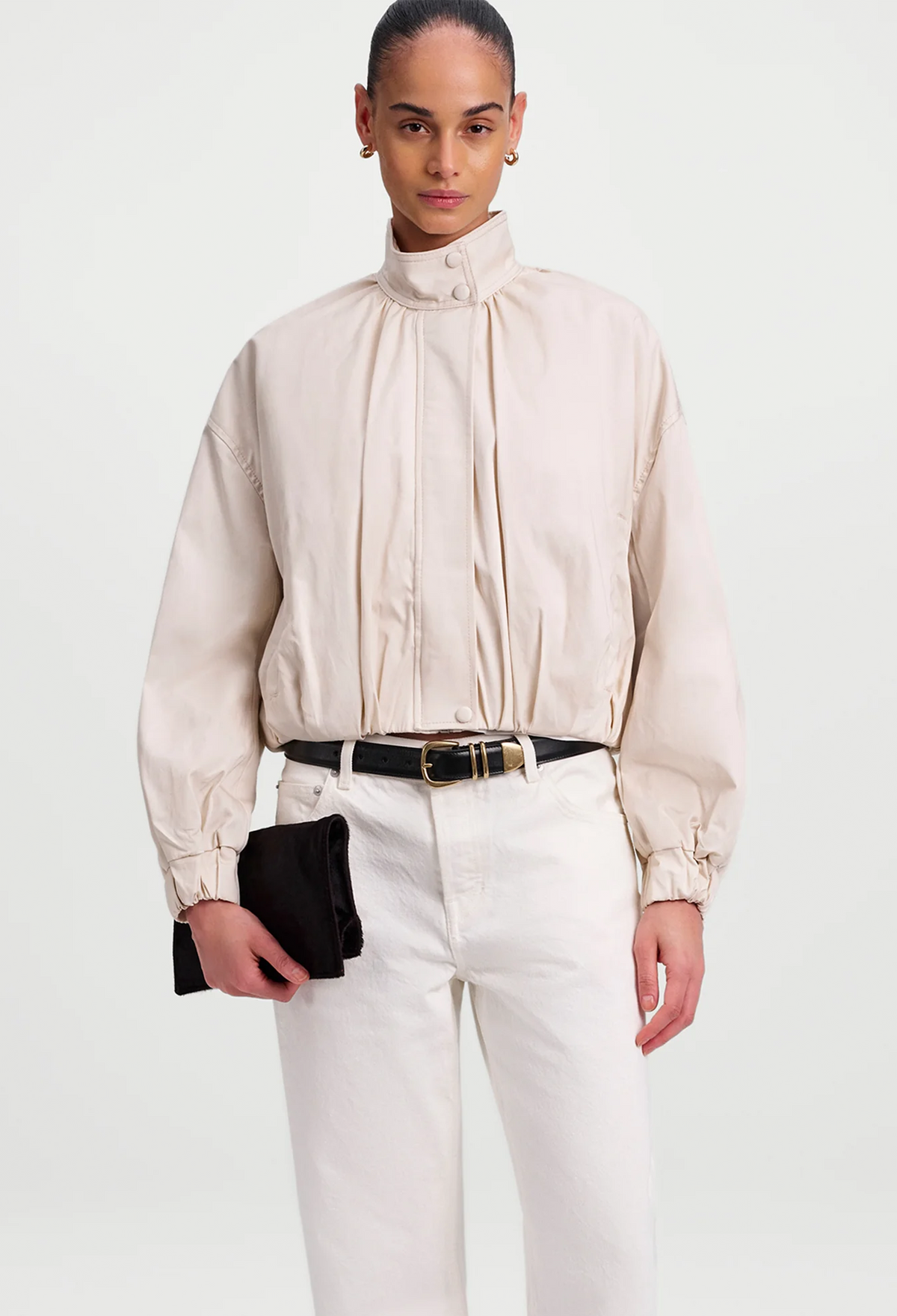 A model wearing a off white high-neck, voluminous long-sleeve jacket with gathered details, paired with white trousers against a light gray background.