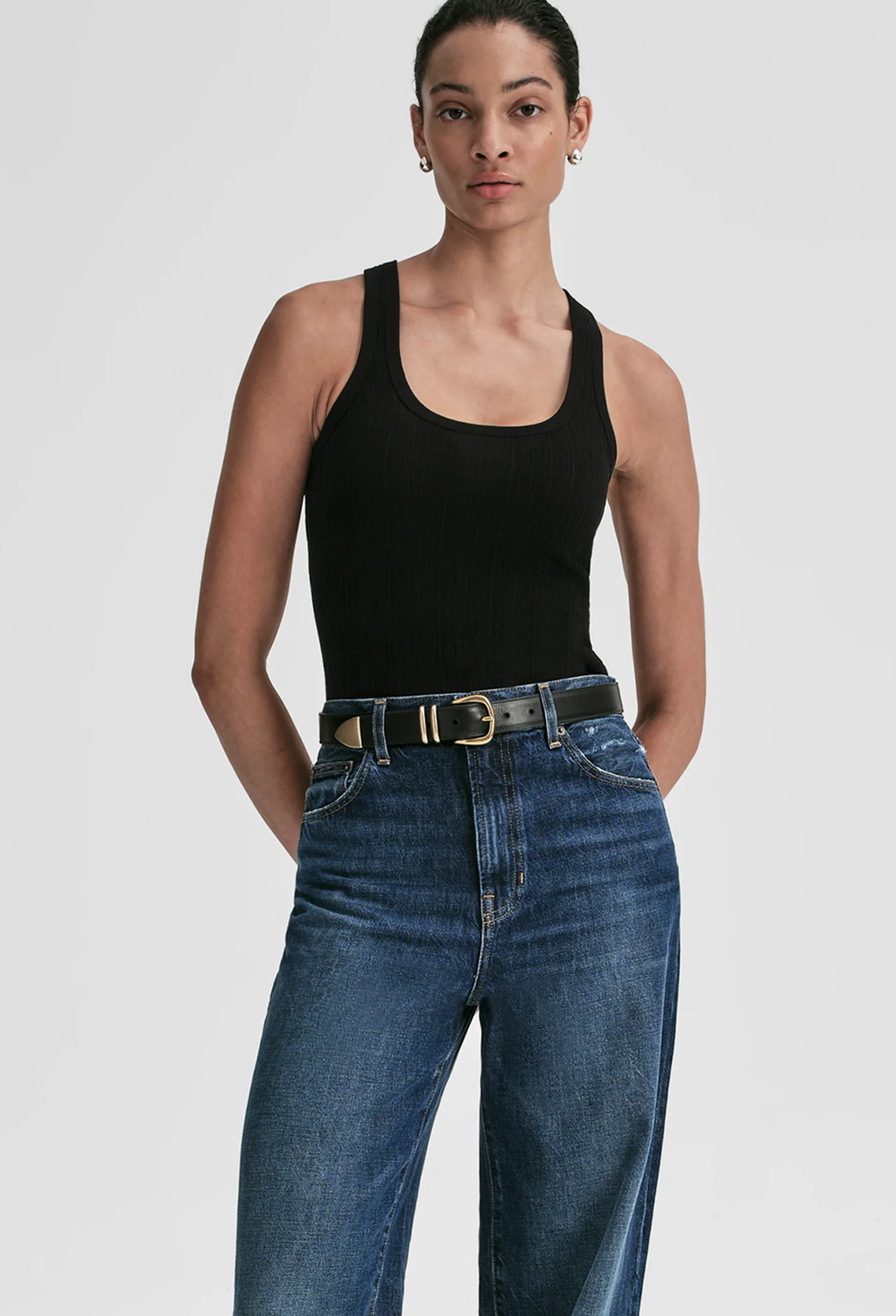 A model wearing a black ribbed tank top and high-waisted blue jeans with a black belt, photographed against a light gray studio background.